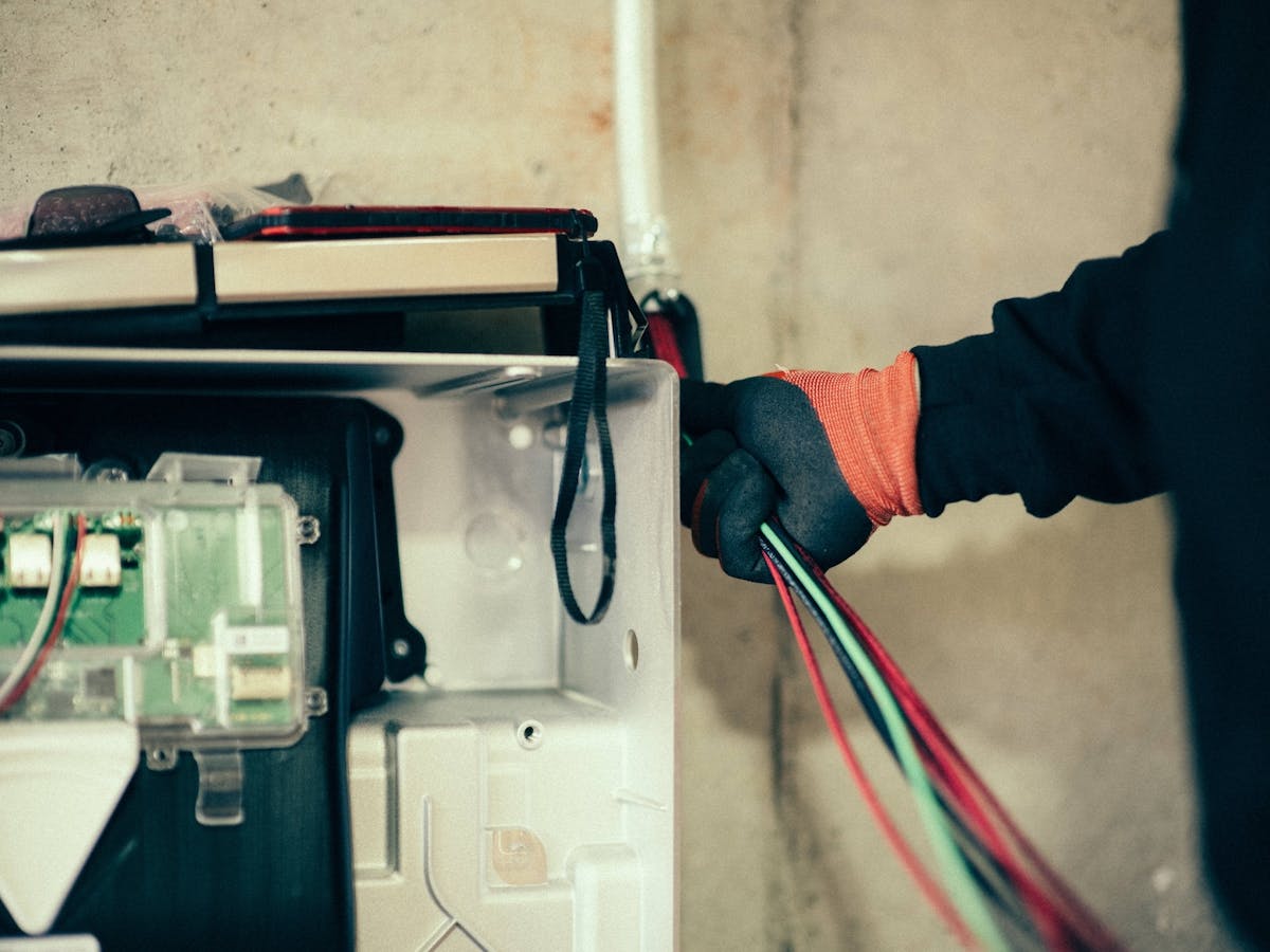 an electrician wearing gloves holds multiple colored wires while working on the internal components of an electrical panel or applianc