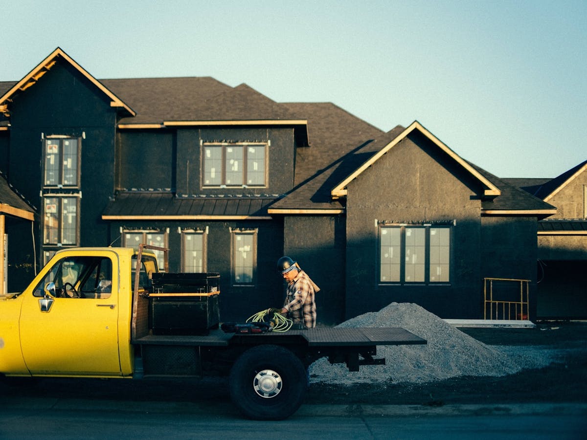 worker stands next to a yellow flatbed truck in front of a house under construction