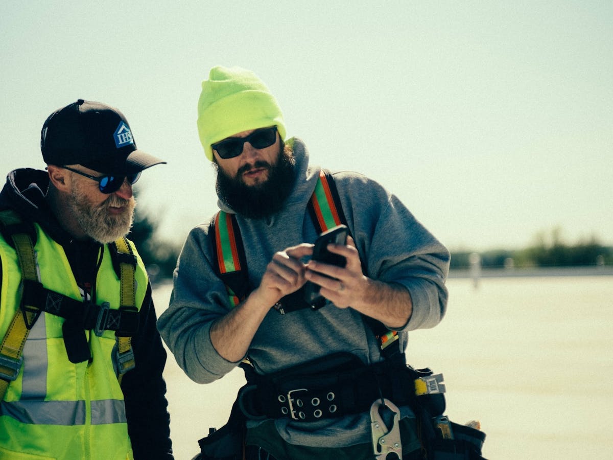 Roofing crew reviews project details on mobile app while wearing harnesses on active rooftop job site.