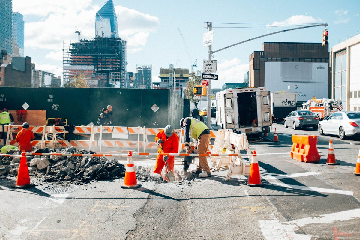 Construction workers jackhammer street pavement inside coned work zone for liability protection.